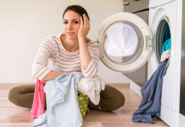 Femme assise par terre devant un lave-linge ouvert avec un panier de linge sale, affichant un air frustré et pensif face à une panne d'appareil.