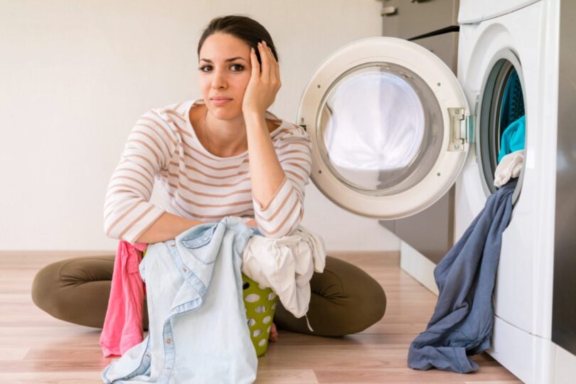 Femme assise par terre devant un lave-linge ouvert avec un panier de linge sale, affichant un air frustré et pensif face à une panne d'appareil.