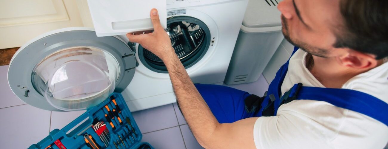 Technicien en salopette bleue réparant un lave-linge avec boîte à outils