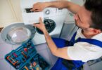 Technicien en salopette bleue réparant un lave-linge avec boîte à outils