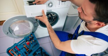 Technicien en salopette bleue réparant un lave-linge avec boîte à outils